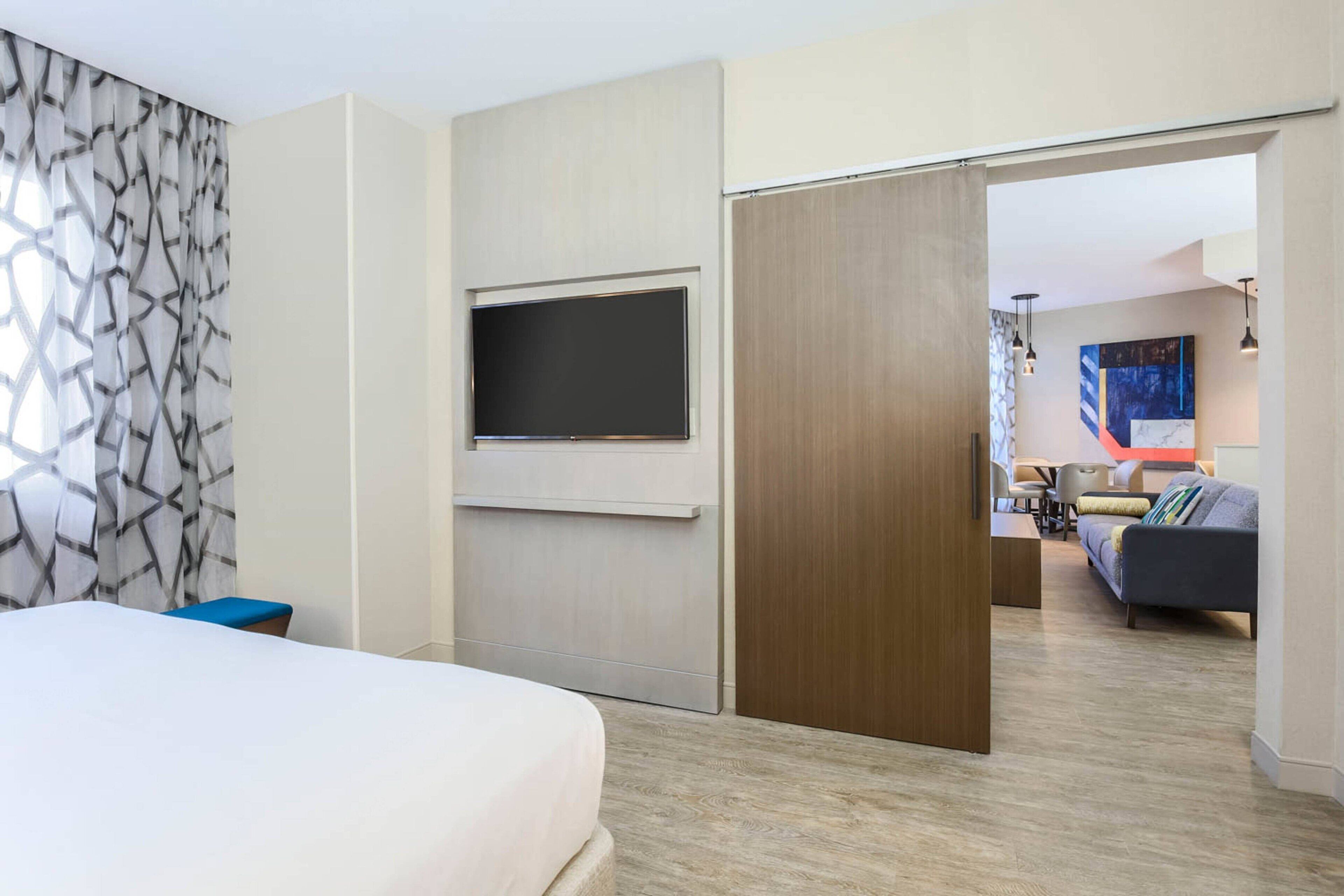 Ein modernes Hotelzimmer mit einem Bett, einem an der Wand befestigten Fernseher und einer Holztür zu einem Wohnbereich mit einer Couch und einem Tisch.