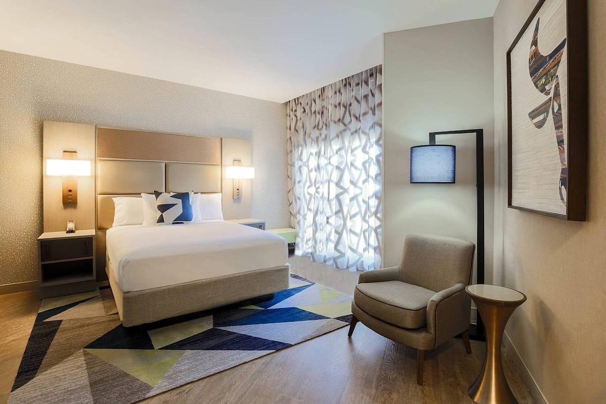 A hotel room with a large bed, bedside lamps, a chair, a small table, and a patterned wall.