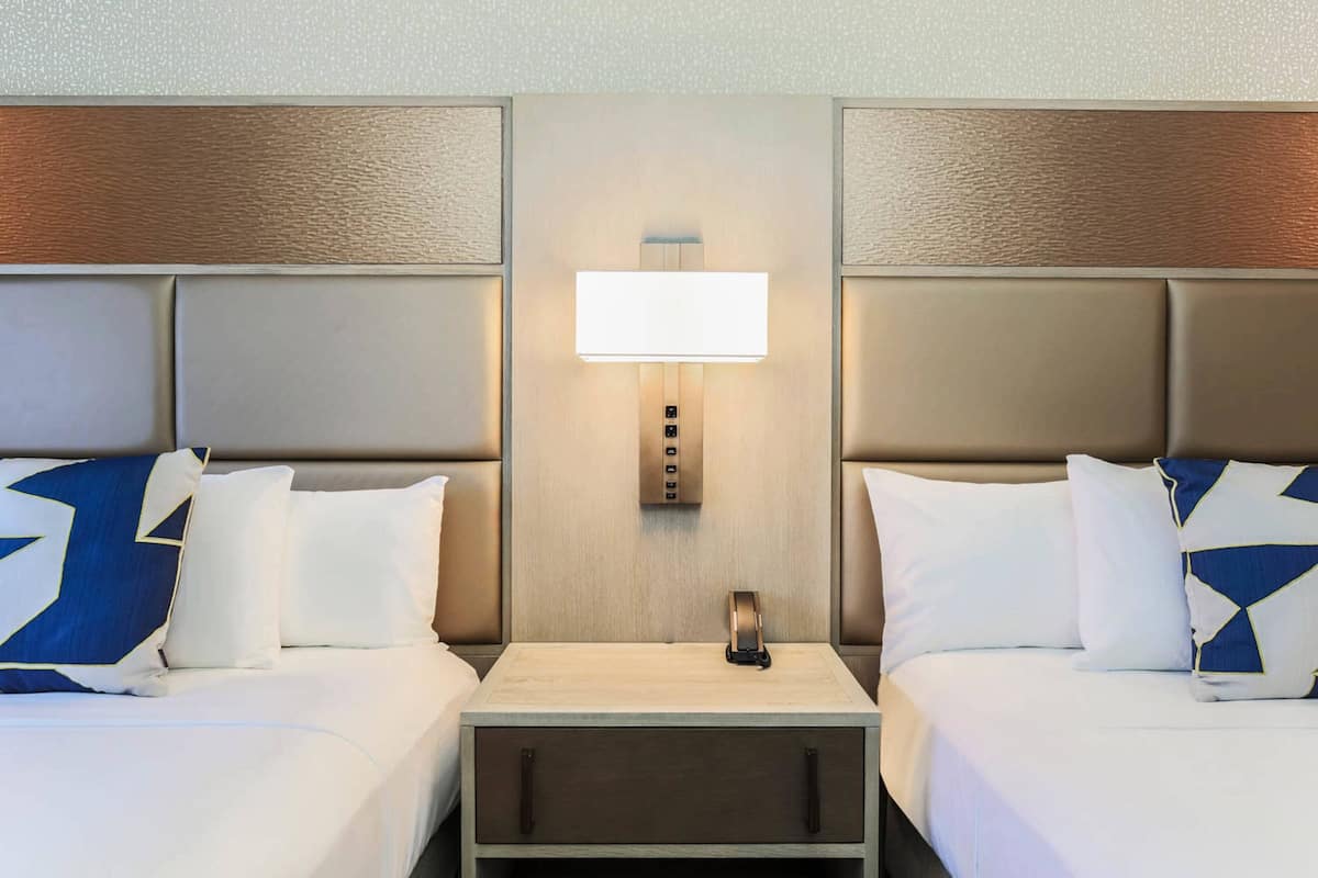 A hotel room with two beds, a nightstand, a wall-mounted lamp, and a small drawer.