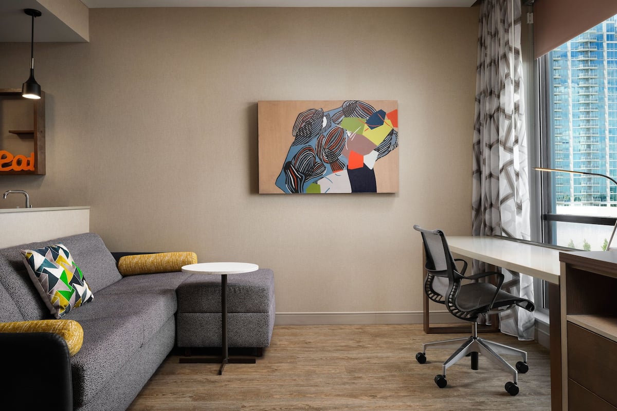 A modern office space with a grey sofa, a round white table, a black office chair, and a colorful abstract painting on the wall.