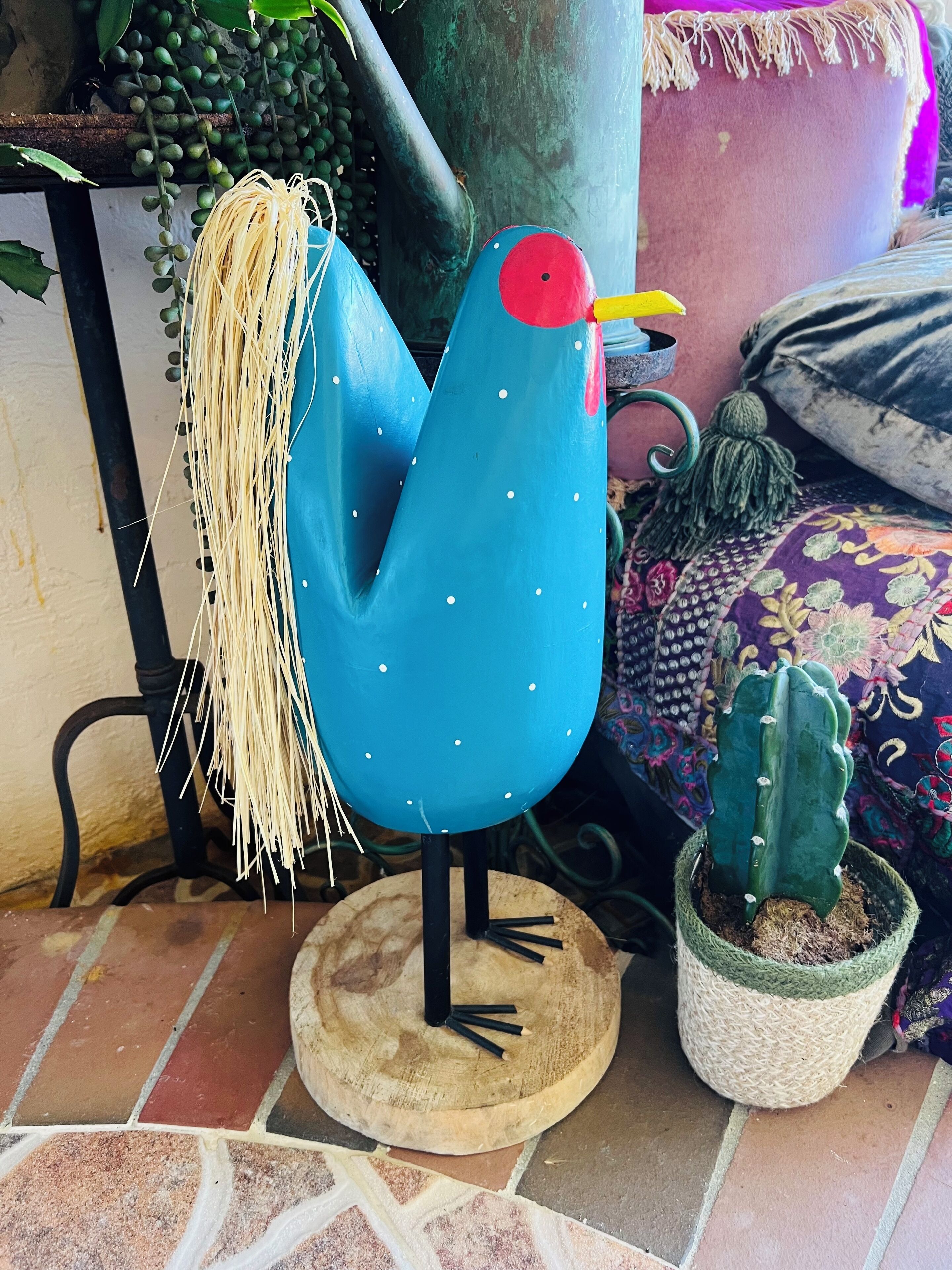 Une chaise bleue en forme d’oiseau, avec une assise en paille, un cactus dans un panier tressé et un coussin aux motifs colorés.