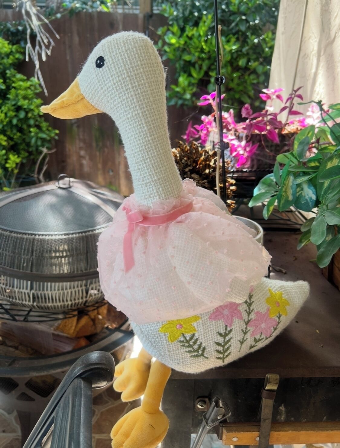 A crocheted duck with a pink bow and floral embroidery, hanging near a grill and plants.