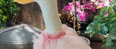 A crocheted duck with a pink bow and floral embroidery, hanging near a grill and plants.