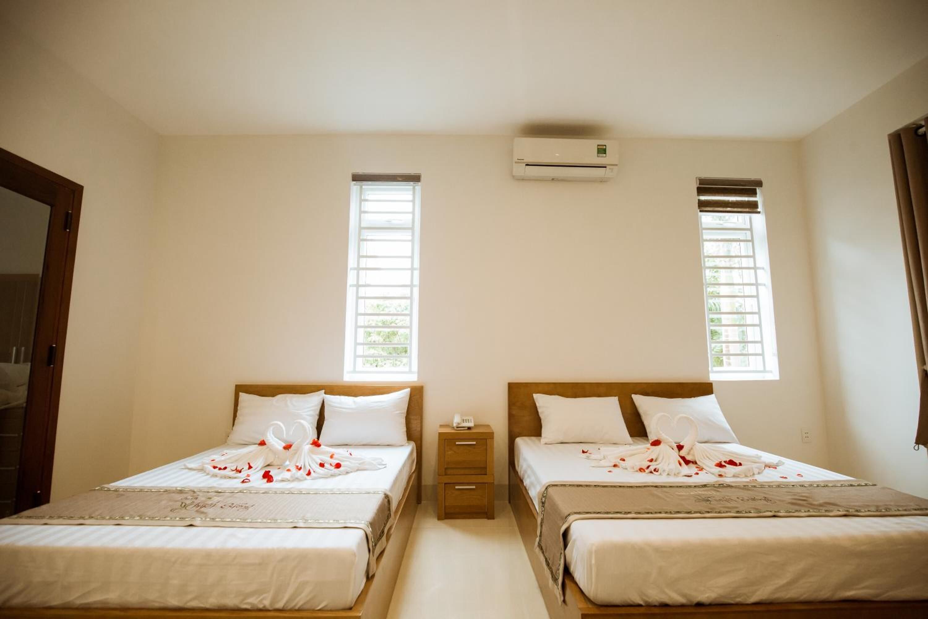 A hotel room with two beds, a small bedside table, an air conditioning unit, and two windows with blinds.