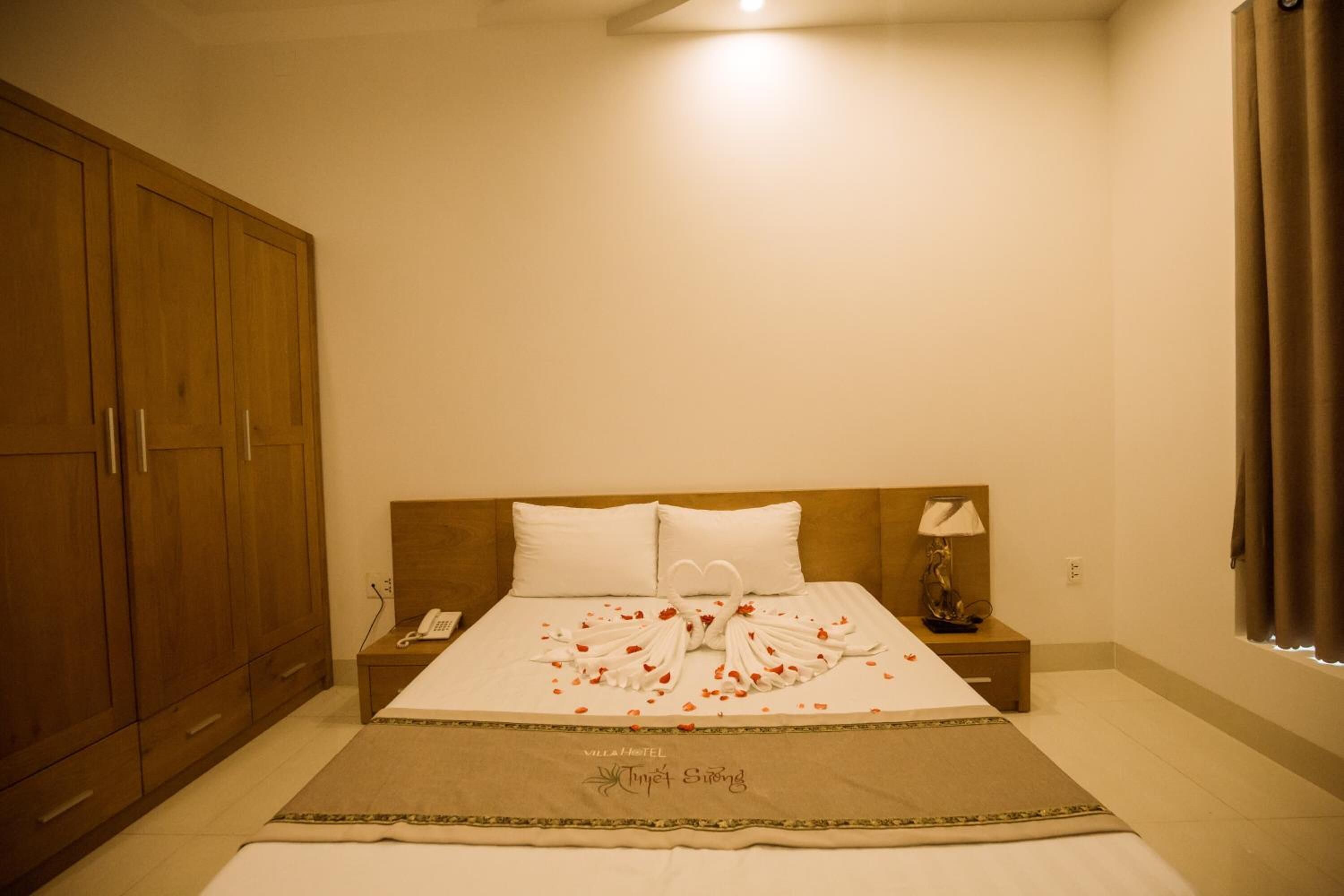 A hotel room with a large wooden wardrobe, a bed with a white bedspread and a decorative swan made of towels, a bedside lamp, and a telephone on the nightstand.