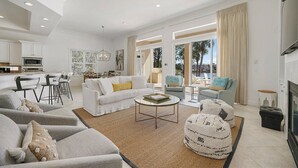 Ein geräumiges Wohnzimmer mit einem weißen Sofa, Sesseln, einem Couchtisch und einem Kamin. Im Hintergrund ist eine Küche mit Essbereich zu sehen.