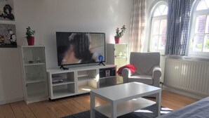 A living room with a flat-screen TV, white furniture, a checkered armchair, and a window with curtains. - Vacation apartment B, Salzgasse, Old Town - Edition Bad Wimpfen (Bad Wimpfen)