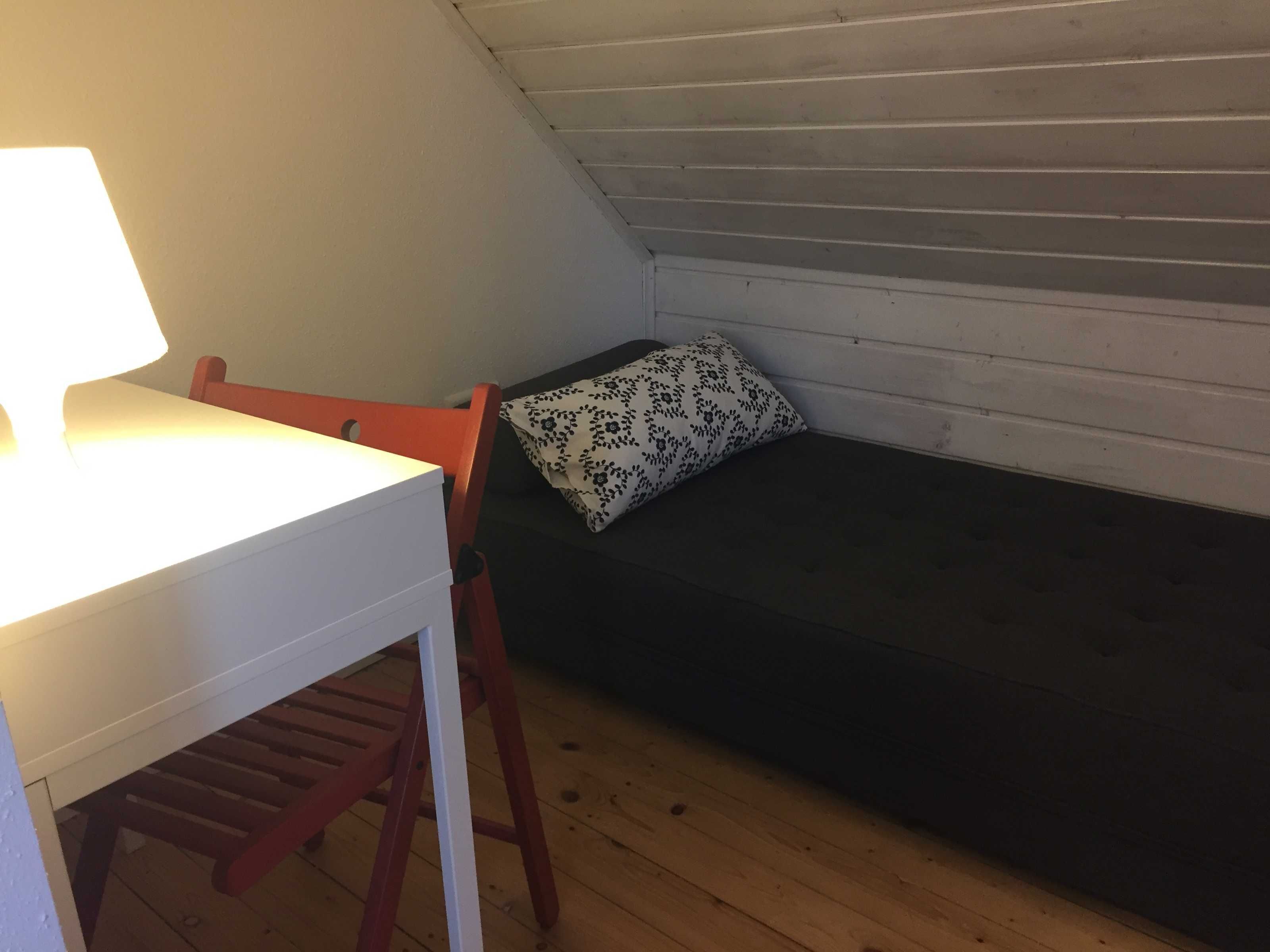 A small, cozy room with a wooden floor, a white desk with a lamp, a red chair, and a dark-colored bed with a patterned pillow.