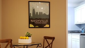 A dining area with a table and chairs, a framed cityscape poster, and a kitchen in the background. - All in Reach, 2BR in West End (Nashville)