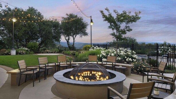 A circular outdoor seating area with a fire pit, surrounded by chairs and tables, set against a backdrop of trees and a view of the landscape. - Hotel Vista Sierra (Auburn)
