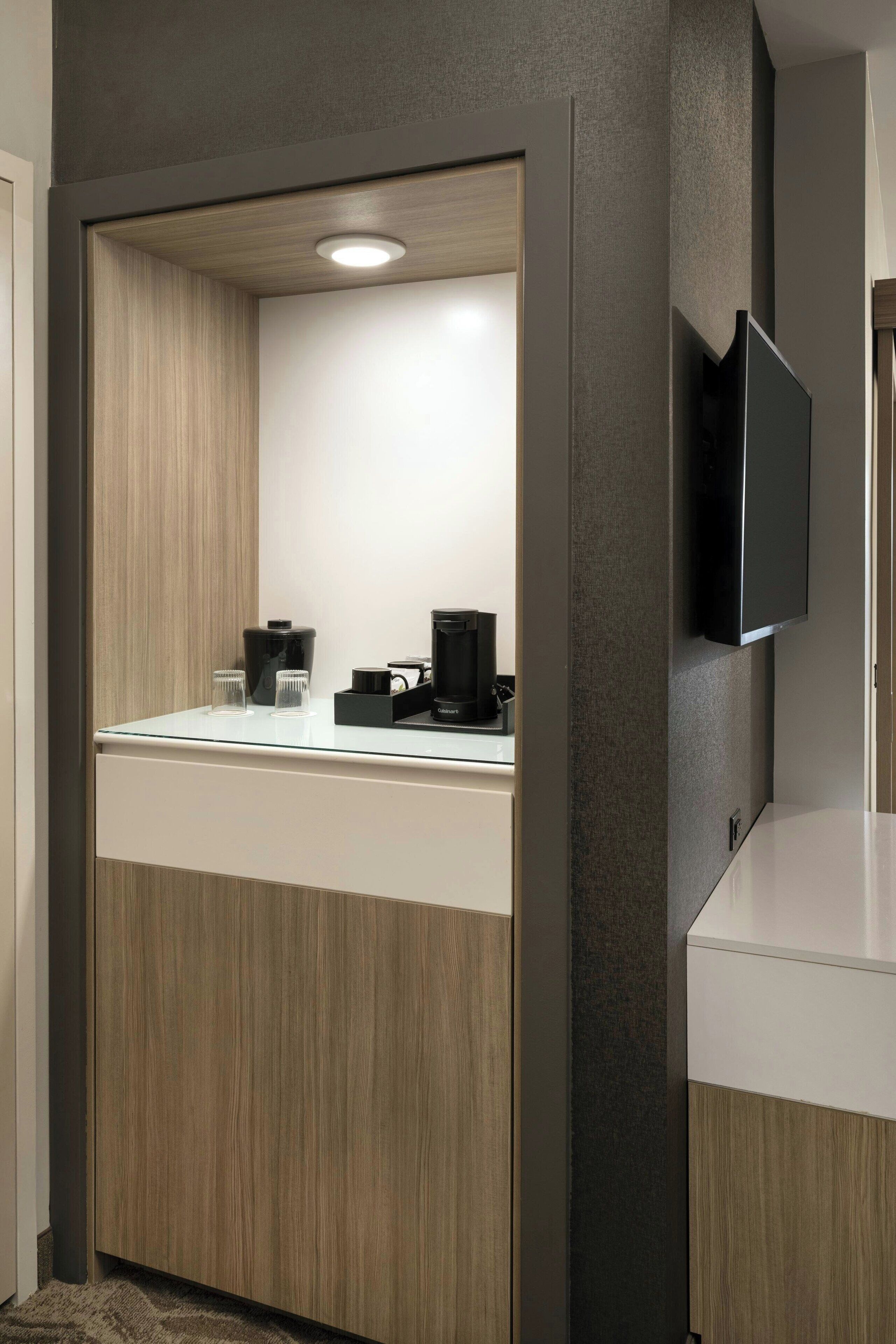 Una habitación de hotel moderna con un tocador de madera, un televisor de pantalla plana montado en la pared y una cafetera en la encimera.