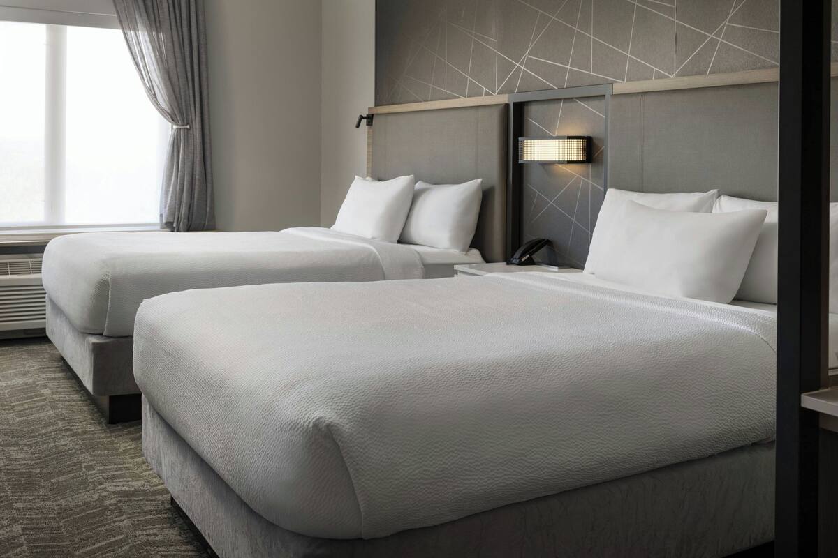 A hotel room with two beds, a modern headboard, a bedside table with a lamp, and a window with curtains.