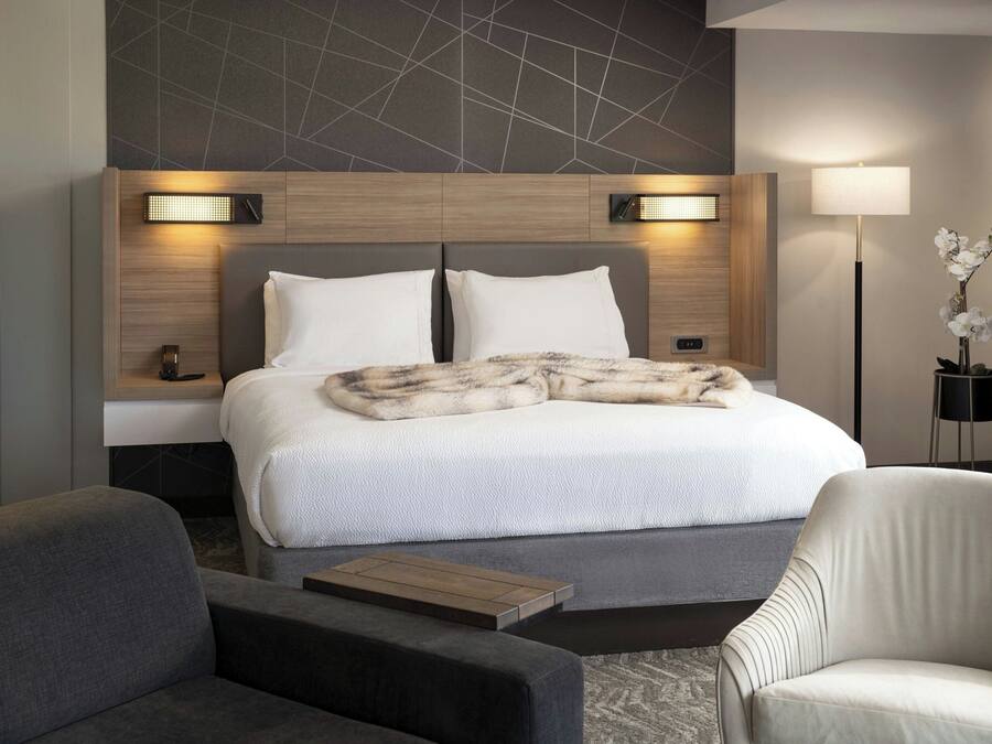 A modern hotel room with a large bed, two bedside tables, a nightstand, a sofa, a chair, and a floor lamp.