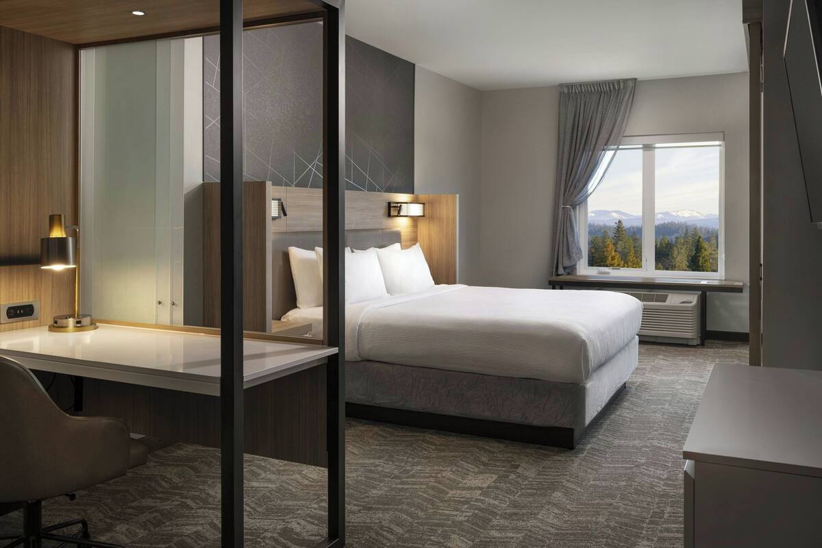 A modern hotel room with a large bed, a desk, a chair, a nightstand, a lamp, a window with a view of mountains, and a sliding door.