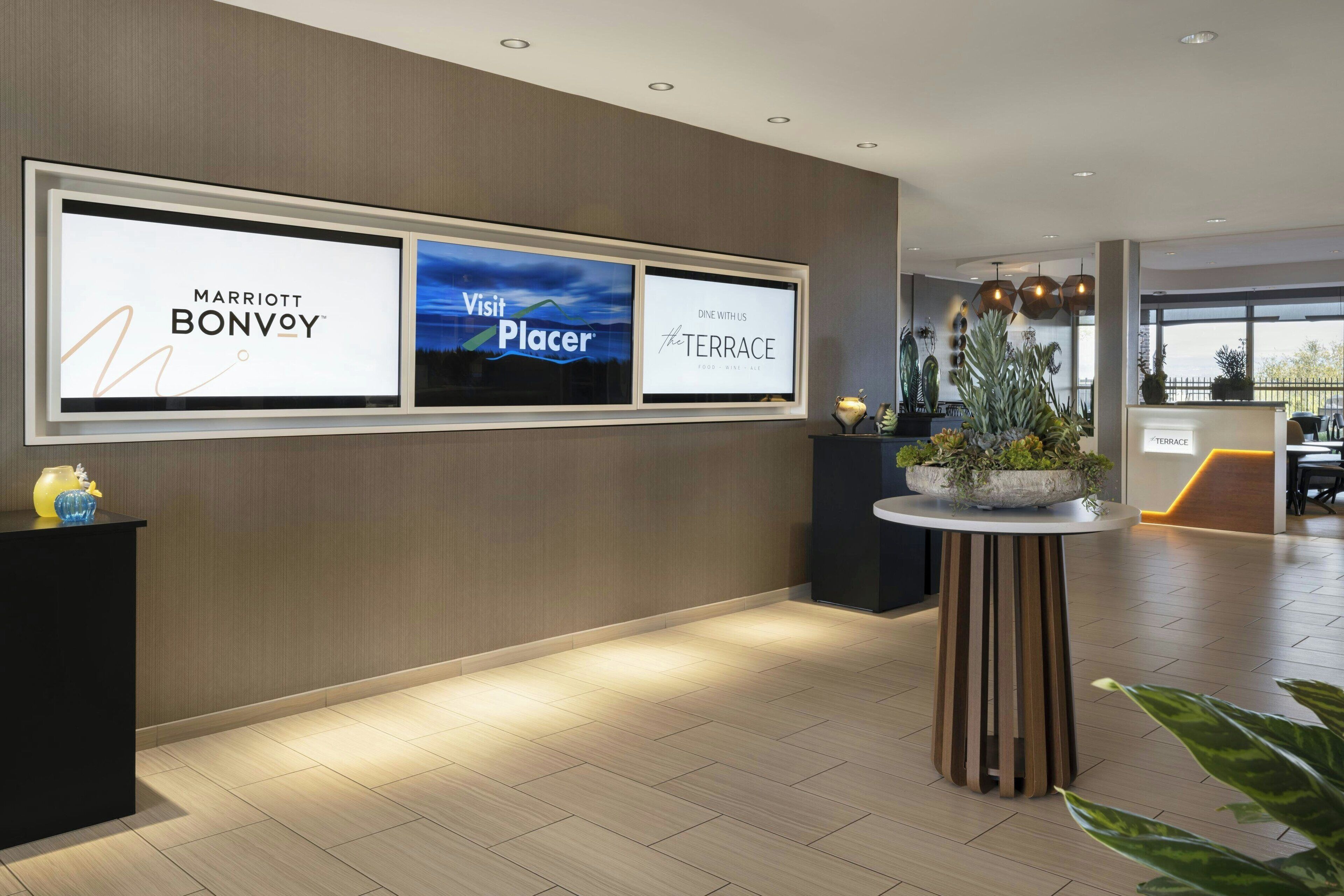 A hotel reception area with a modern design, featuring a reception desk, a digital display board with hotel names, and a potted plant.