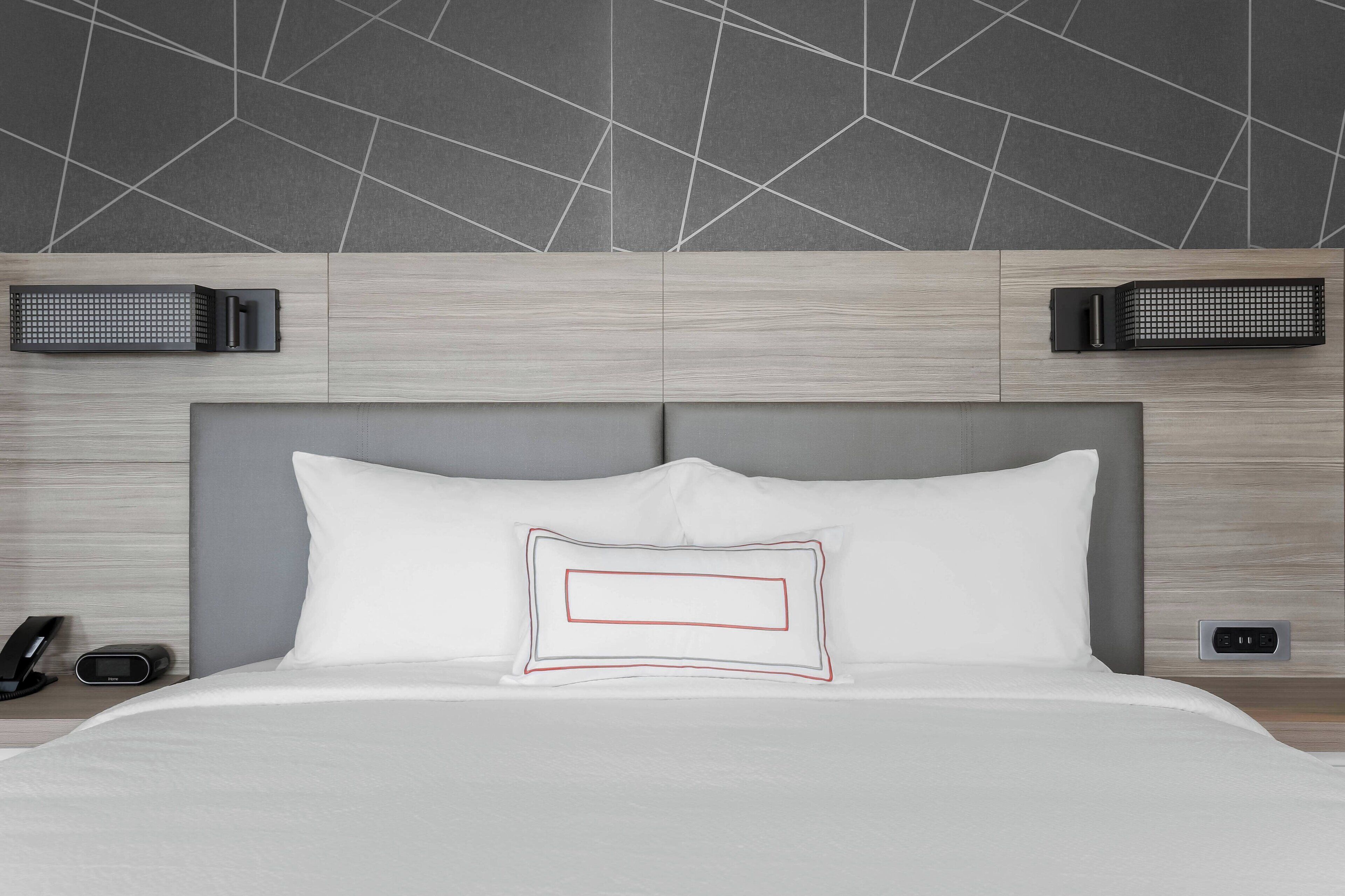 Une chambre d’hôtel moderne dotée d’un grand lit, d’une tête de lit avec éclairage intégré, d’un mur orné d’un motif géométrique et d’une table de chevet avec un téléphone et une horloge numérique.