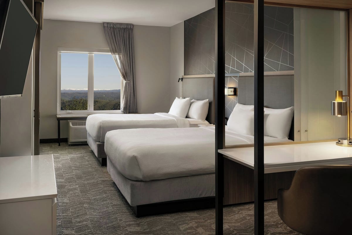A hotel room with two beds, a desk, a chair, and a large window offering a view of the landscape.