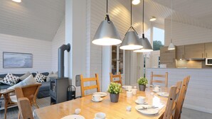 A modern dining area with a wooden table set for four, surrounded by wooden chairs. There are pendant lights above the table, and a kitchen area is visible in the background.