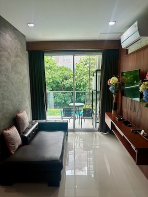 A modern living room with a sofa, a flat-screen TV, and a balcony with a view of greenery. - Pool view 1BR Apartment +Pool Access by PW (Hua Hin)