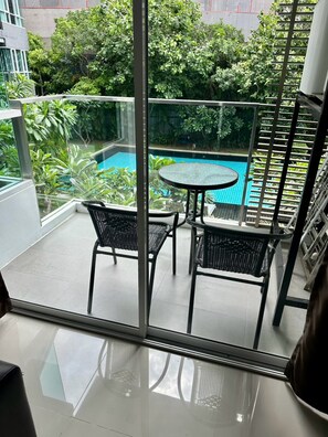 A balcony with a glass partition, a small round table, two chairs, and a view of a swimming pool surrounded by greenery.