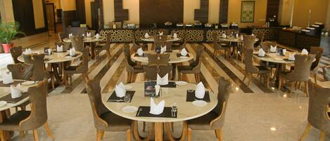 A spacious hotel dining area with multiple round tables and chairs, a reception desk, and a large plant.