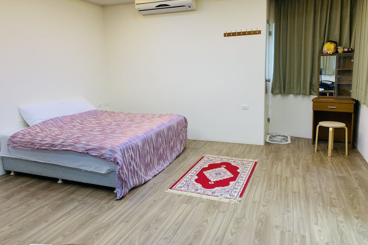A bedroom with a bed, a wooden desk, a chair, a small rug, a mirror, and an air conditioning unit.