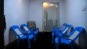 Quarto com quatro cadeiras de plástico azul dispostas em semicírculo, uma pequena mesa com uma moldura e uma janela com cortinas translúcidas.