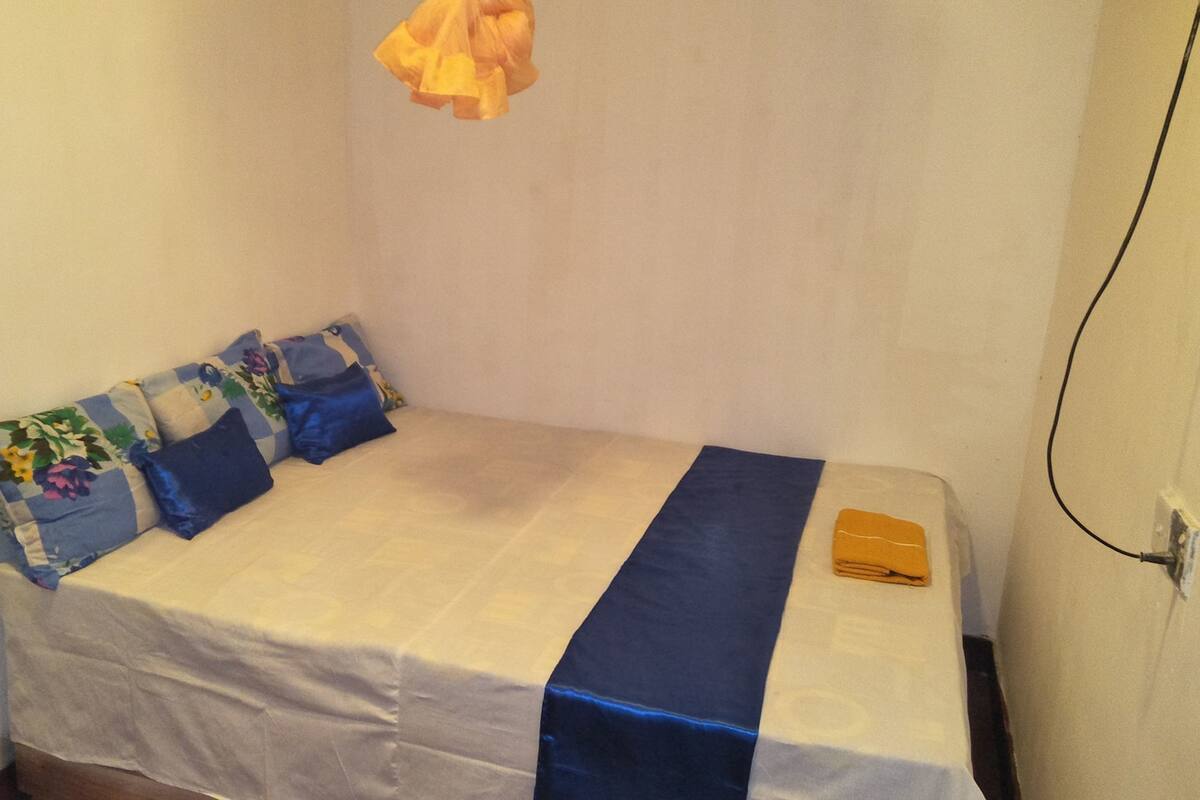 A small, single-bed room with a white and blue striped bedspread, several pillows, and a folded towel on the bed.