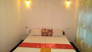 Uma cama de solteiro com colcha vermelha e branca, duas almofadas brancas e uma almofada acolchoada colorida. Há duas luminárias fixadas na parede e uma tomada.