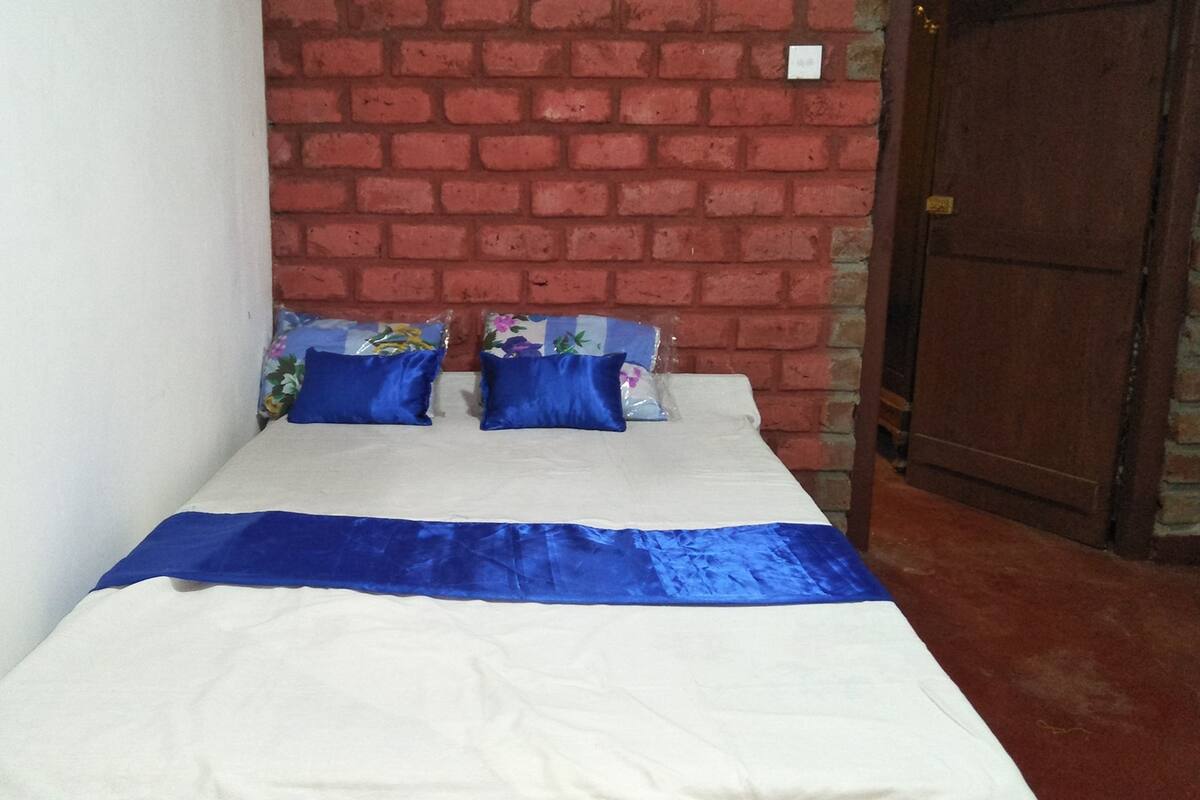 A single bed with a white and blue bedspread, two blue pillows, and floral-patterned pillows against a brick wall.