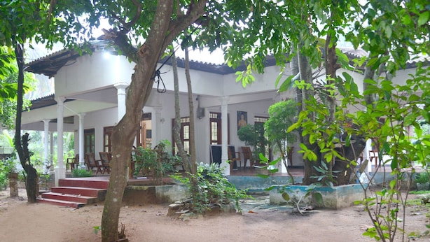 Um edifício tradicional com varanda coberta, degraus que levam a uma porta e um pequeno jardim com plantas e uma árvore.