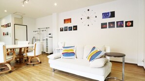 A modern living room with a white sofa, a dining table, and a small round table. There are wall-mounted pictures and a unique clock. - 3A Spacious room with Built-in Kitchen (Ōsaka-shi)