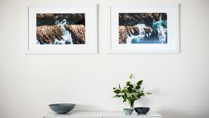 Zwei gerahmte Fotografien einer felsigen Küstenlinie mit Wellen, ein weißer Konsolentisch mit einer Schale und einer Pflanze sowie zwei Stühle davor.