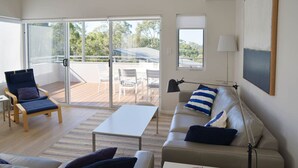 Ein modernes Wohnzimmer mit Sofa, Sessel und Couchtisch. Ein großes Schiebeglastür führt zu einer Terrasse mit Sitzgelegenheiten.
