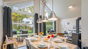 Spiseområde med et bord dækket til fire personer, store vinduer og et moderne køkken i baggrunden.