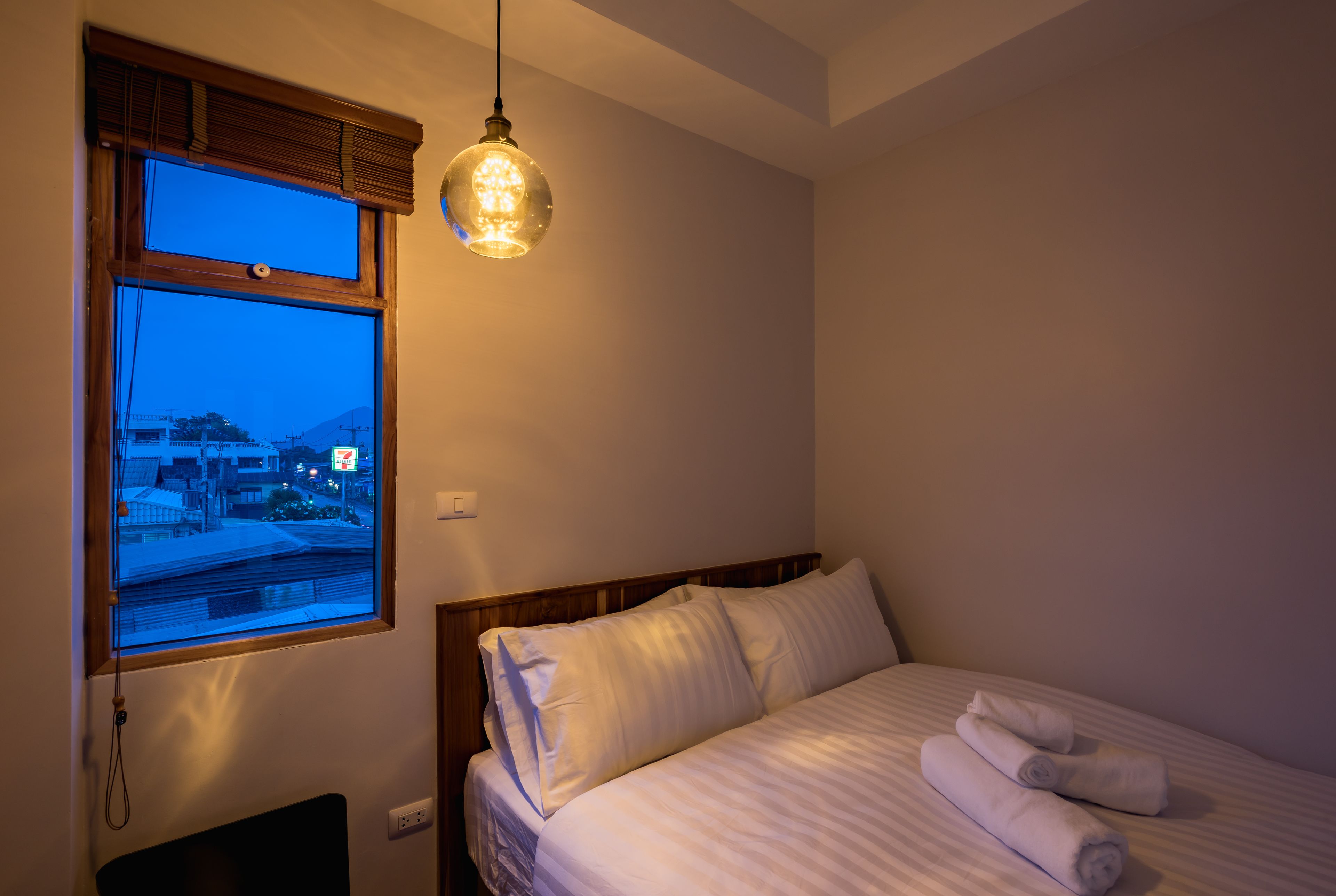 A bedroom with a bed, pillows, a bedside table, and a window with a view of buildings and mountains.
