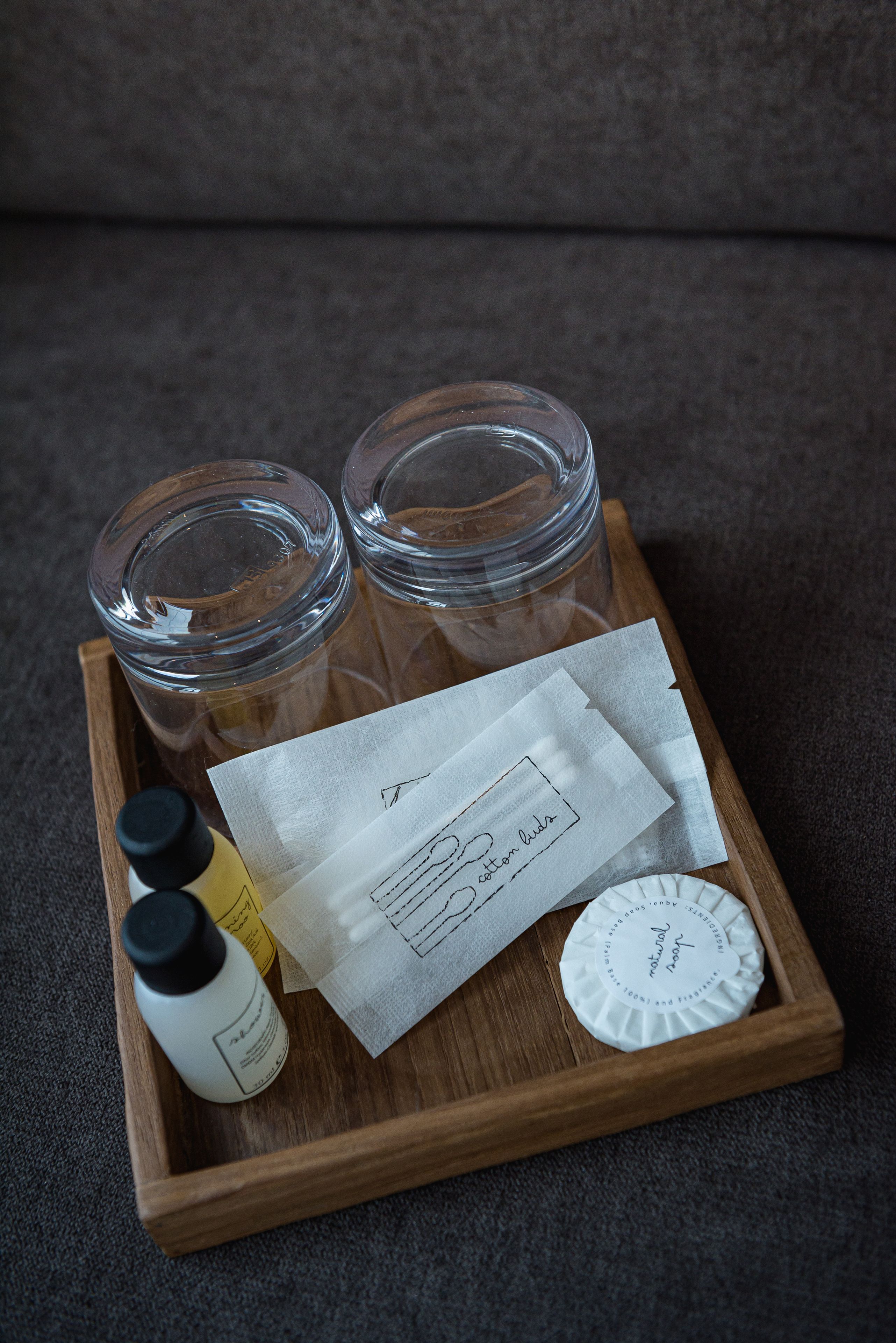 Un plateau en bois contenant deux bocaux en verre, une bouteille blanche, un mouchoir, un petit récipient blanc et une feuille de papier avec du texte.