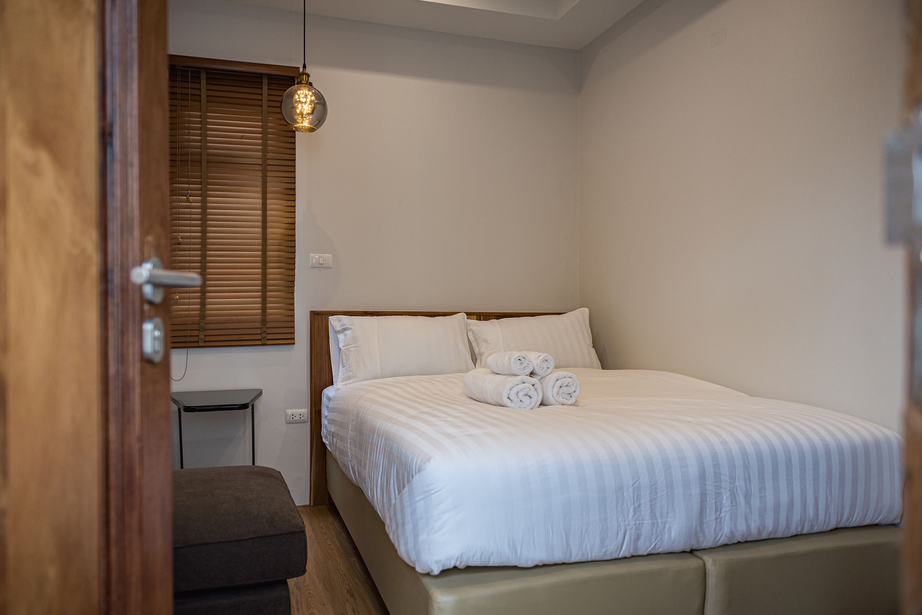 A bedroom with a bed, a small table, a bench, and a window with blinds.