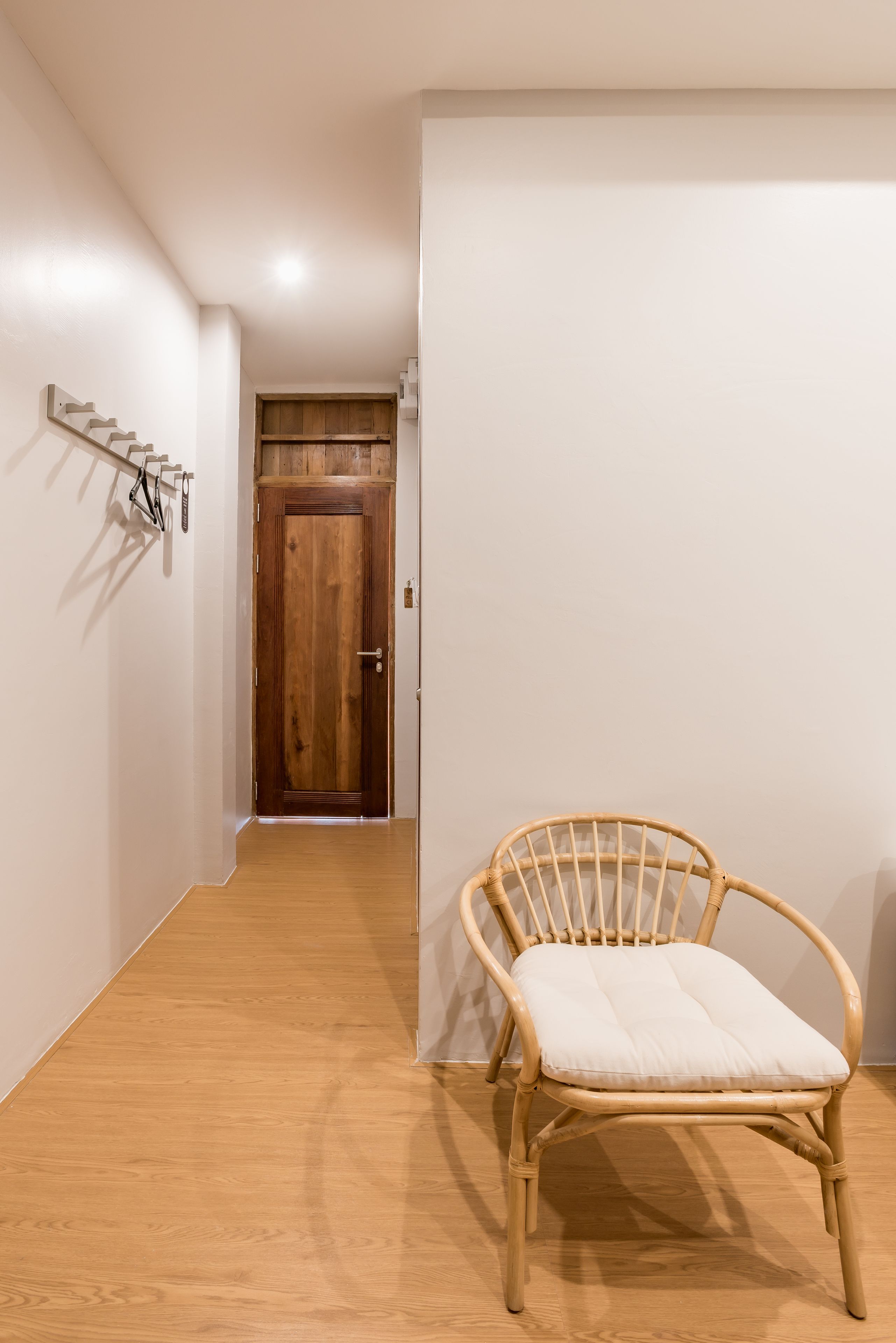 A hallway with a wooden door at the end, a wicker chair with a cushion, and a coat rack with hangers.