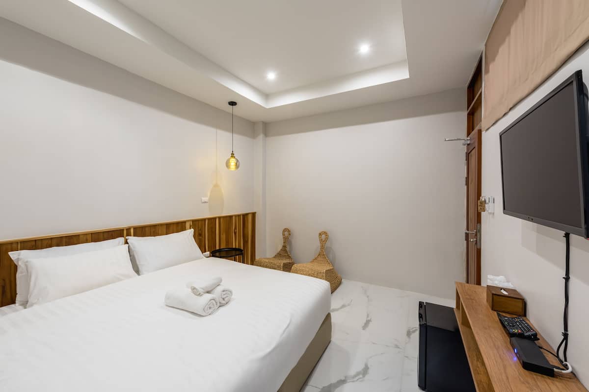 Quarto de hotel moderno com uma cama bem arrumada, cabeceira de madeira, TV de tela plana na parede e uma pequena escrivaninha com computador e teclado.