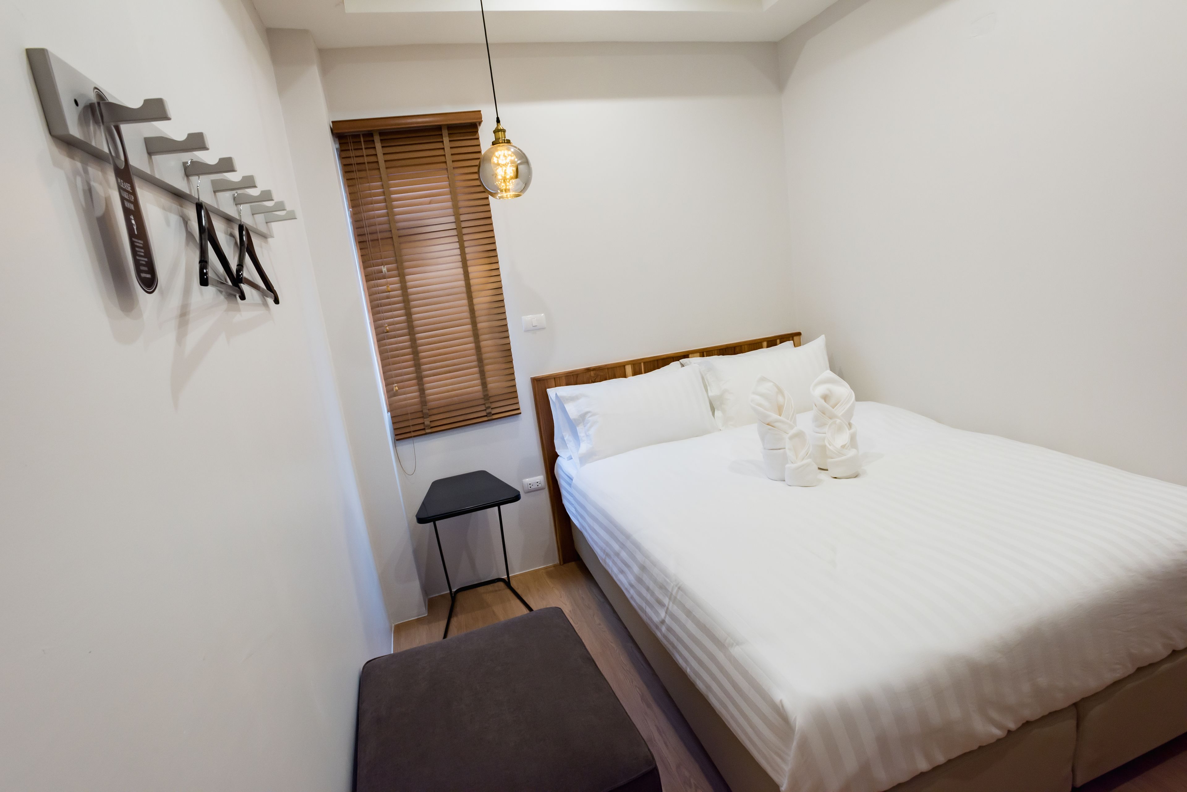 Un dormitorio con cama, un banco, una mesita, un perchero de pared y una ventana con persianas.