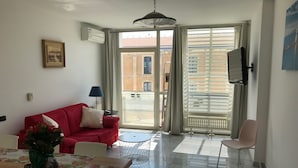 A living room with a red sofa, a dining table with a vase of flowers, a TV mounted on the wall, and a large window with blinds.
