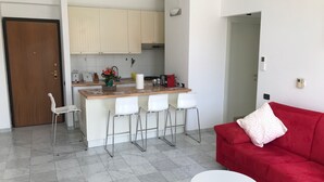 A modern kitchen with white cabinets and a central island with bar stools. There is a red sofa and a white round table in the living area.