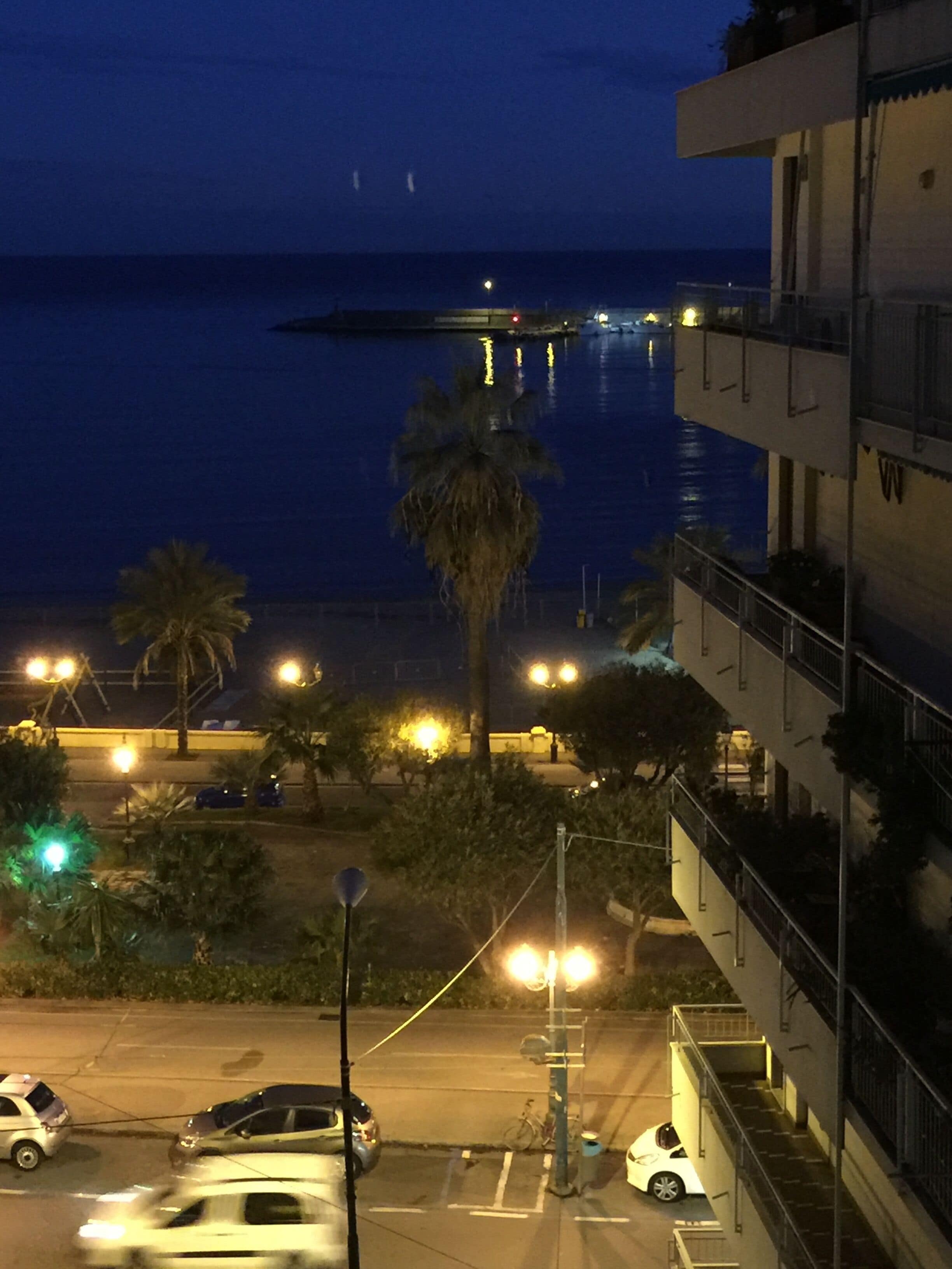 A nighttime view of a coastal area with a building overlooking the sea, street lights, and parked cars.