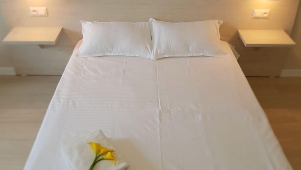 A neatly made bed with white linens, two pillows, and a folded towel with a yellow flower on a wooden floor. - Sky Hotel (Bishkek)
