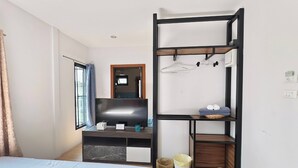 A hotel room with a bed, a TV, a desk with a computer, a shelf with clothes and towels, and a window with curtains. - Lilliput Riverside Bangkachao (Phra Pradaeng)