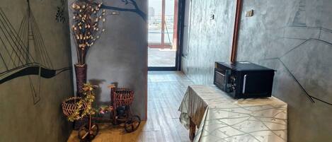 Una habitación con suelo de madera, una mesa con mantel estampado, un jarrón decorativo con ramas y una pared con un mural de un árbol y un puente.