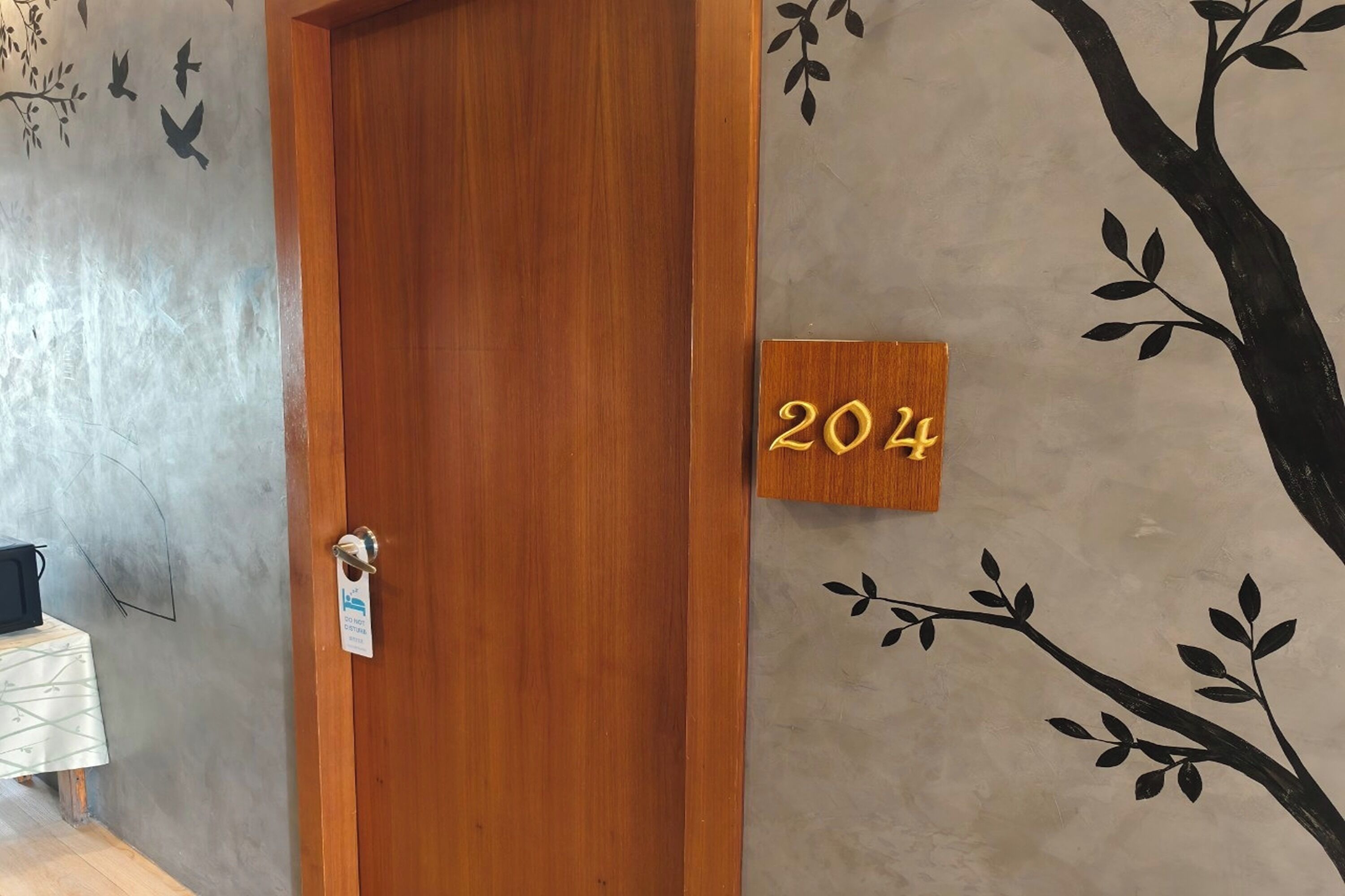 Une porte en bois portant une plaque avec le numéro « 204 » et un mur orné d’une fresque représentant une branche d’arbre.