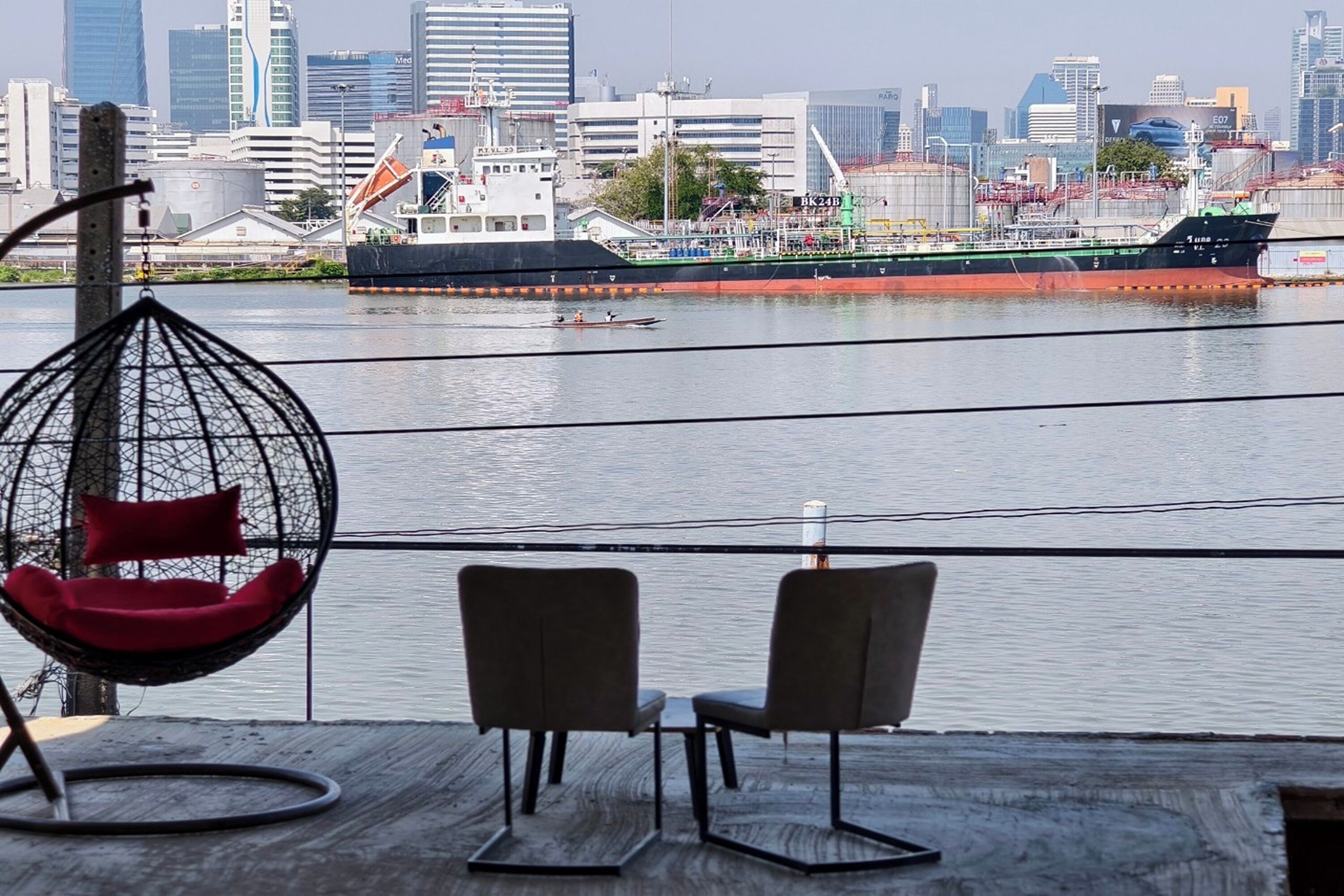 A waterfront view with a city skyline, a large ship, and a seating area with chairs and a hanging chair.