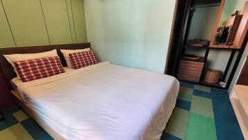 Cama com roupa de cama branca e almofadas xadrez, chão quadriculado e um espelho sobre uma prateleira de madeira com objetos.
